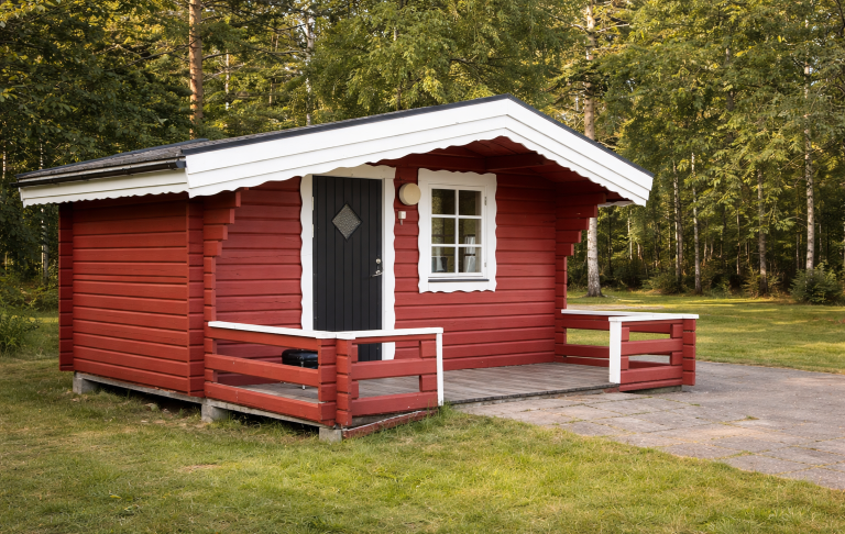 Röd stuga med vit kant, veranda och grönskande omgivning.