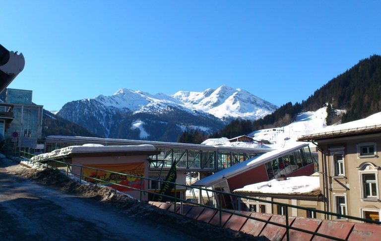 Snöklädd bergskedja med granar och en frusen sjö i förgrunden. Klara blå himmel.