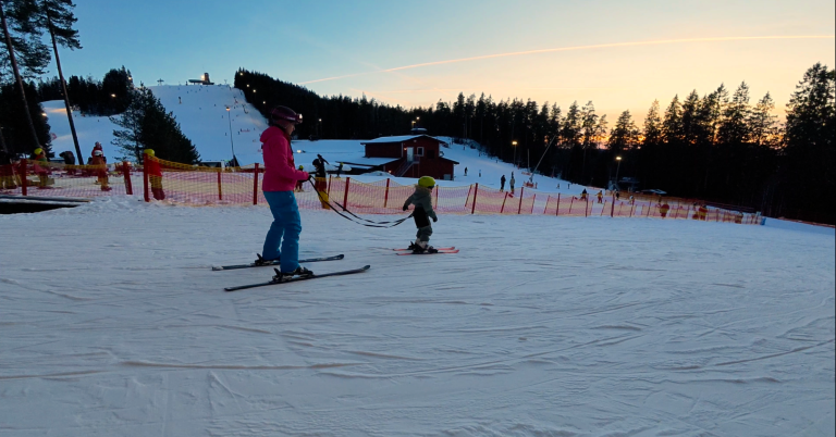 En vuxen och ett barn åker skidor på snöklädda backar vid solnedgång.