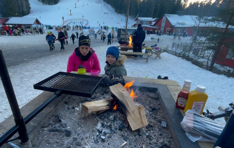 En vuxen och ett barn grillar vid en öppen eld på snöig plats med röda stugor i bakgrunden.