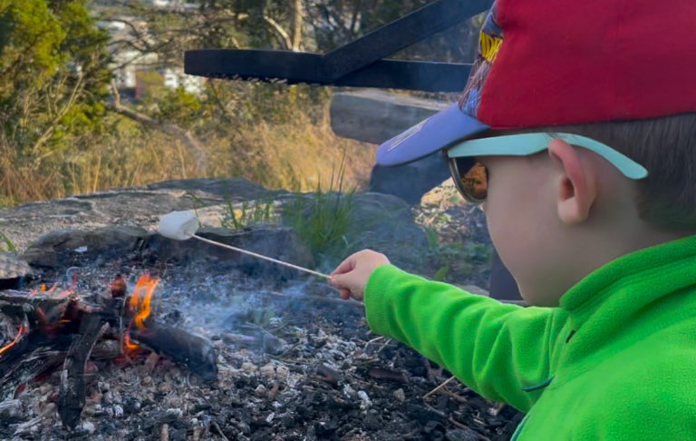Ett barn i grön jacka lagar mat över en öppen eld.