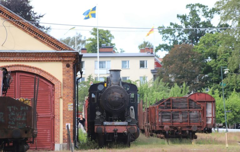 Ånglok framför en tågdepå med vagnar, omgiven av träd och byggnader.