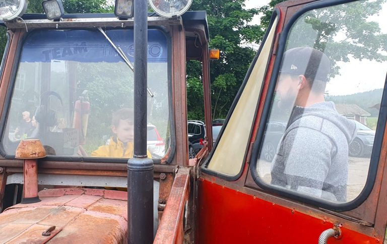 Barn sitter i en traktor medan en vuxen står intill, omgivna av grönska.