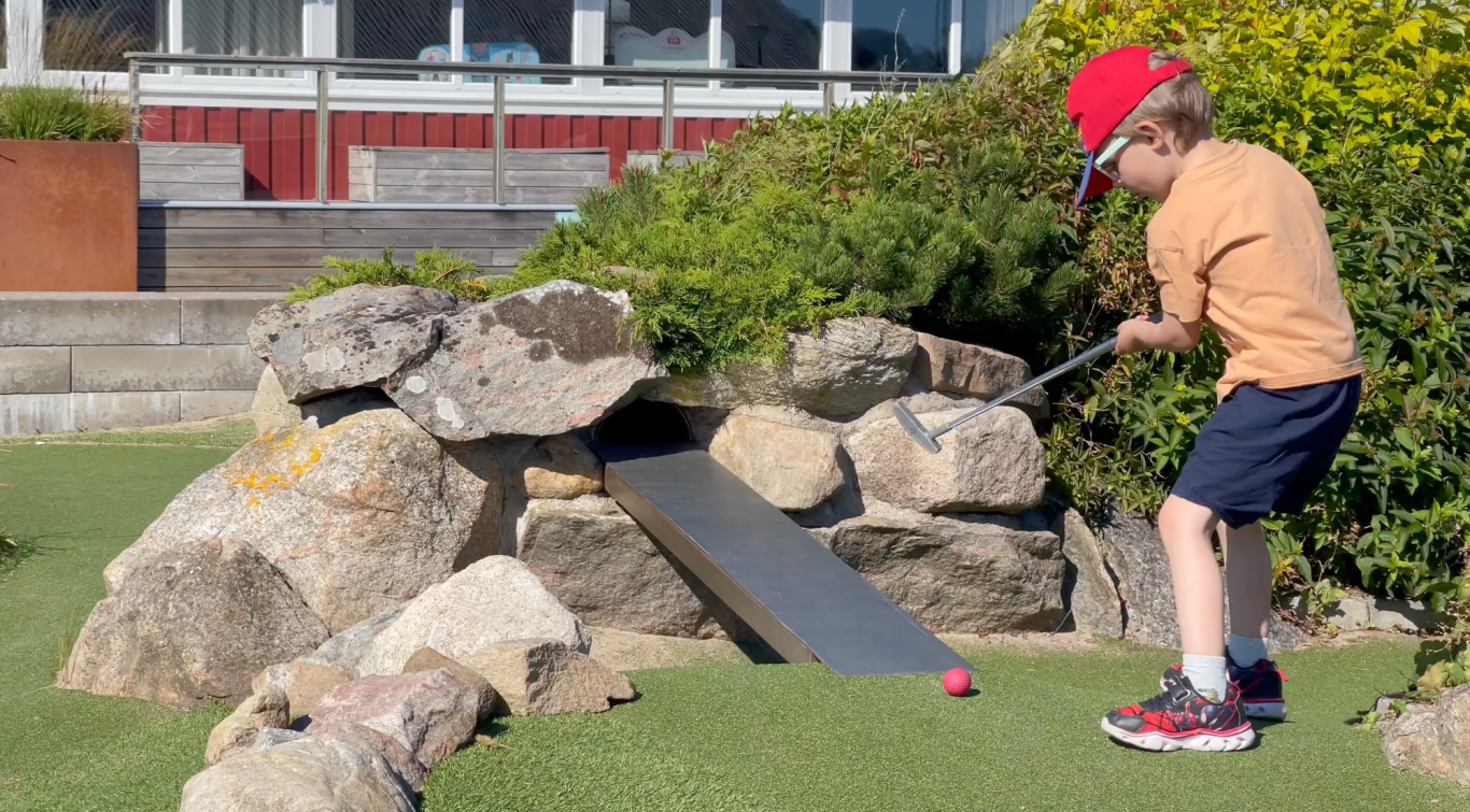 Läjets Camping Äventyrsgolf Ett barn spelar minigolf med en putter nära en stenig bana.