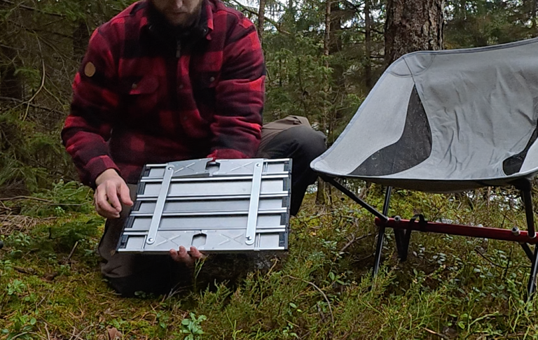 Person i skog visar en ihopvikbar campingstol och en platta med cylindriska delar.