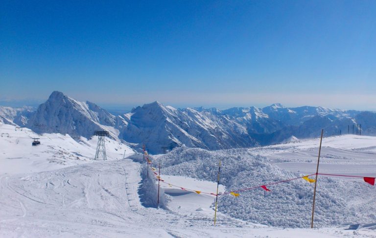 Snöklädda alptoppar med klarblå himmel och en färgglad pistpinne i förgrunden. Skidbacke.