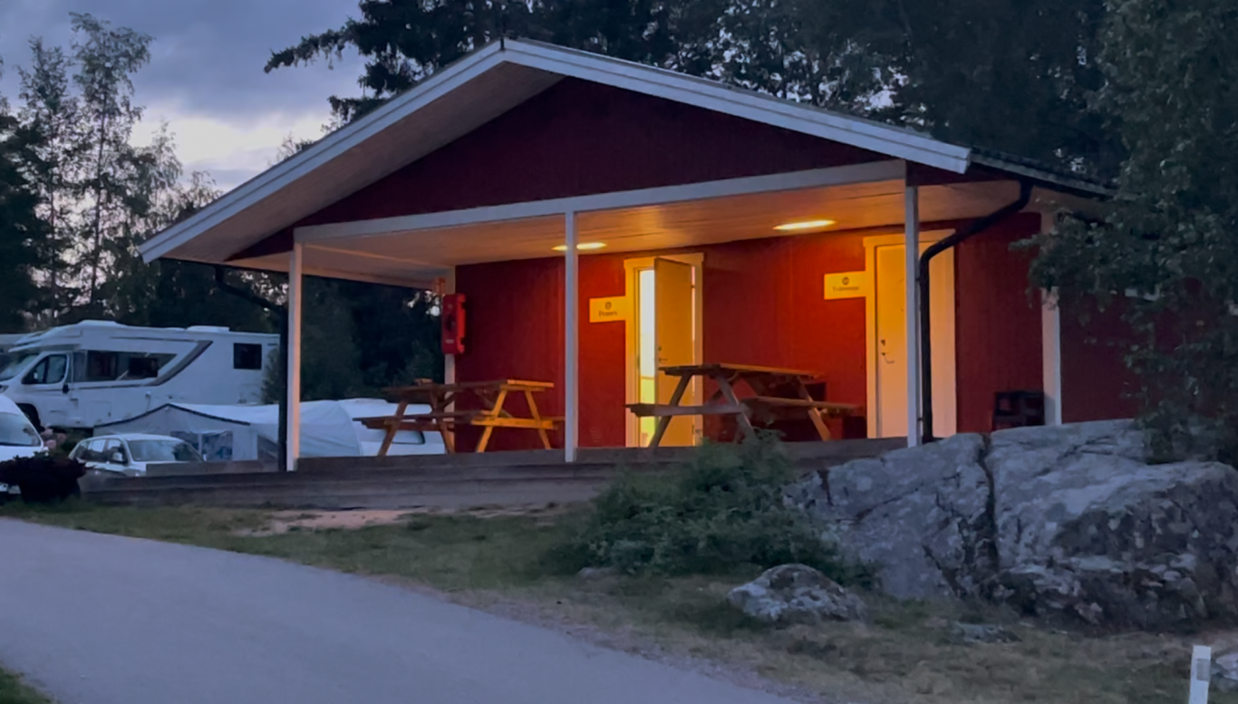 Diskrummet Röd stuga med upplysta fönster och bänkar, omgiven av skog och grusväg.