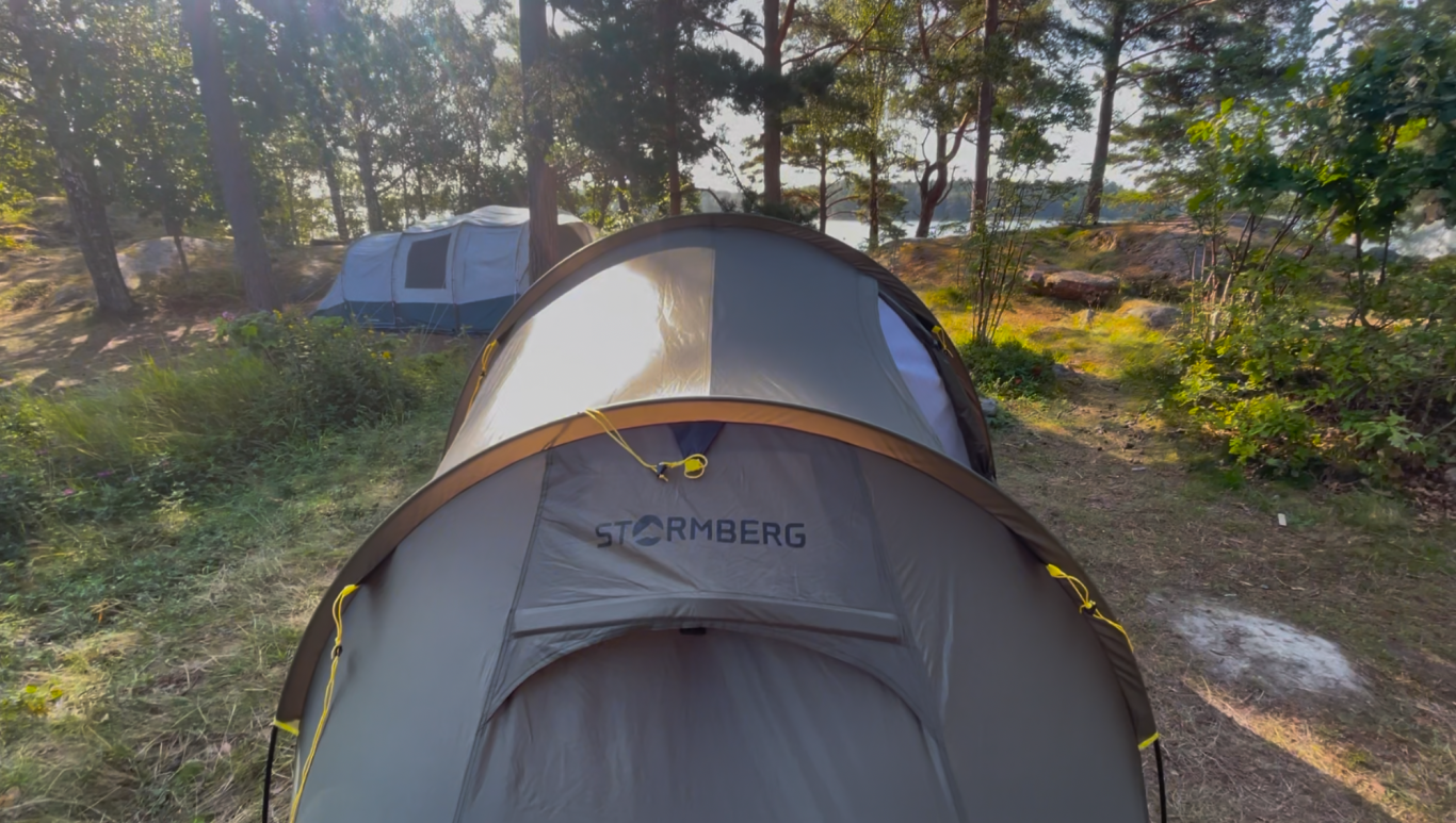Vårt tält Tält i en skog, med en annan tält synlig i bakgrunden och sjö i fjärran.