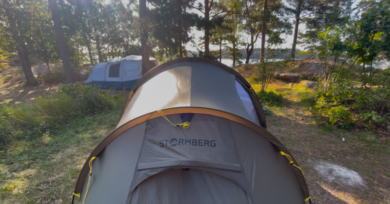 Tält i naturen, omgiven av träd och en annan campingutrustning i bakgrunden.