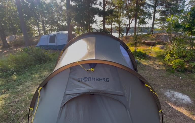 Två tält på en campingplats omgiven av träd och en sjö i bakgrunden.