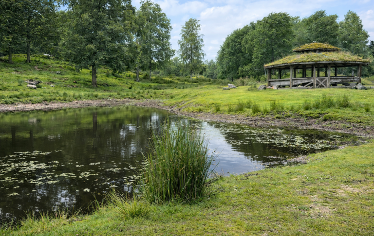 Lummig park med en damm och en paviljong omgiven av grönska.