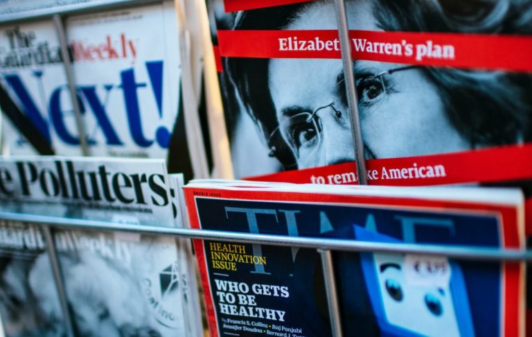 Tidningar med olika omslag, inklusive "Next" och "TIME", staplade i en tidningshållare.