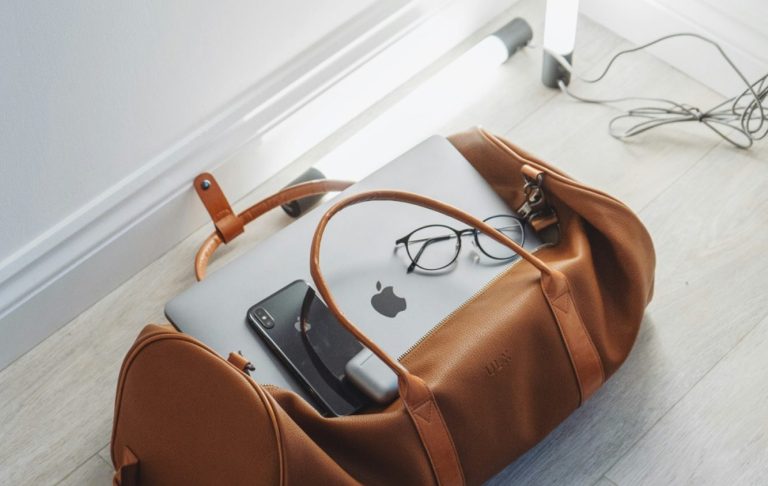 En brun väska med en laptop, mobil och glasögon på ett golv.