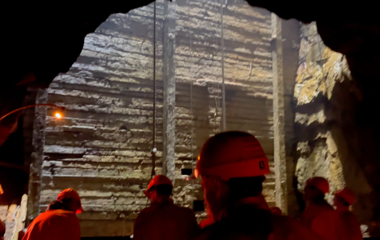 Gruvarbetare i hjälmar står i en tunnel med väggar av berg och belysning.