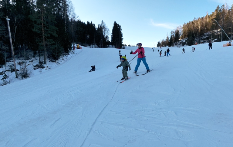 En vuxen hjälper ett barn att åka skidor på en snötäckt backe.