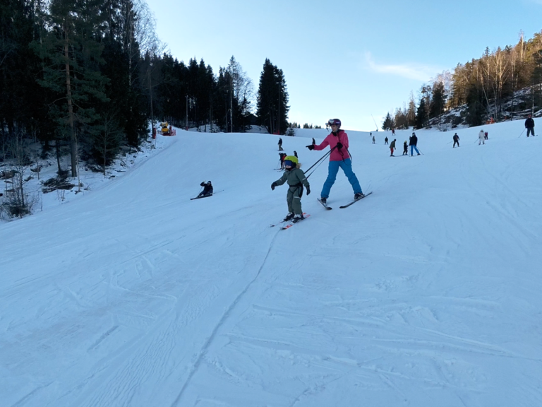 Två skidåkare på snötäckt backe, en vuxen som hjälper ett barn att åka.