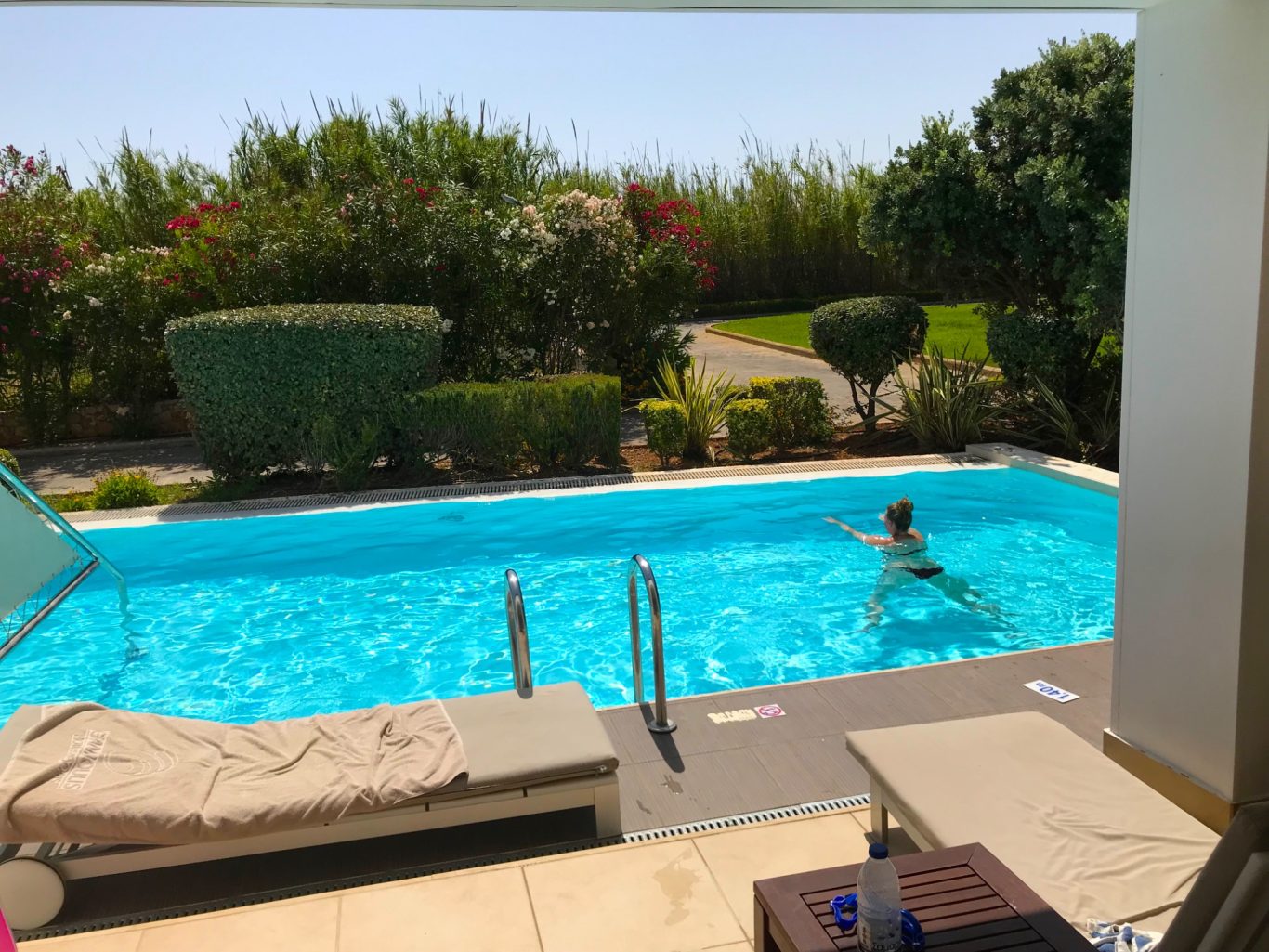 Rosita på Simmtur En person simmar i en blå pool omgiven av grönska och soliga omgivningar.