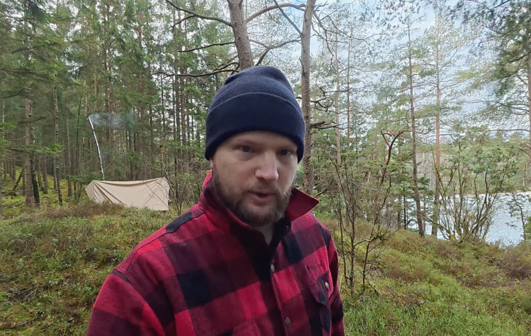 Mann i rutig jacka och mössa stående vid en skogssjö.