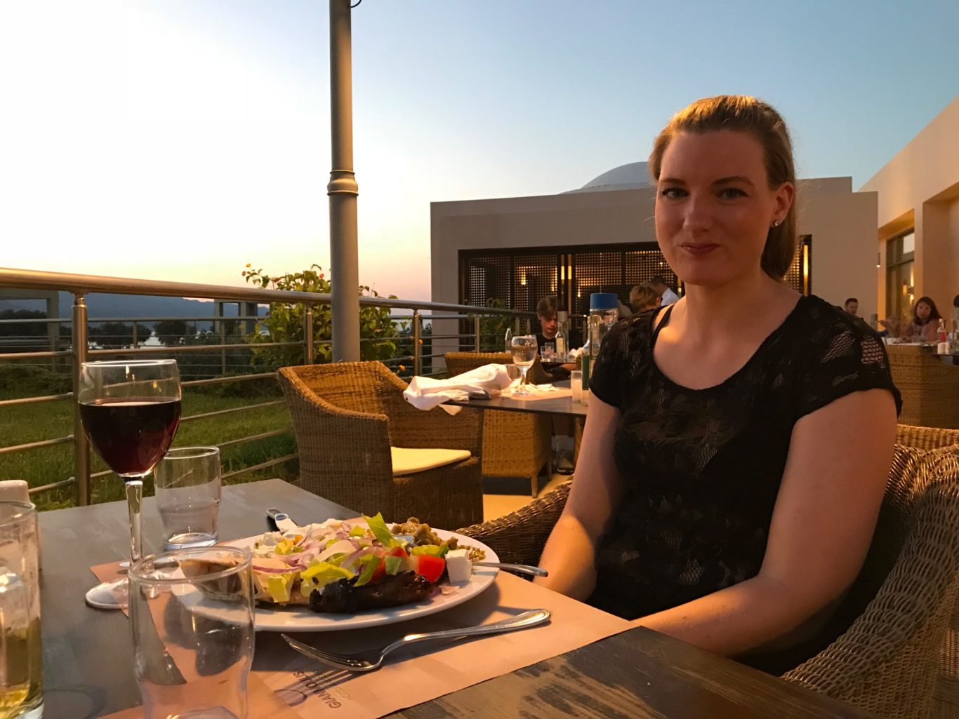 Middag på Buffen Kvinna sitter vid ett bord med mat och vin, solnedgång i bakgrunden.