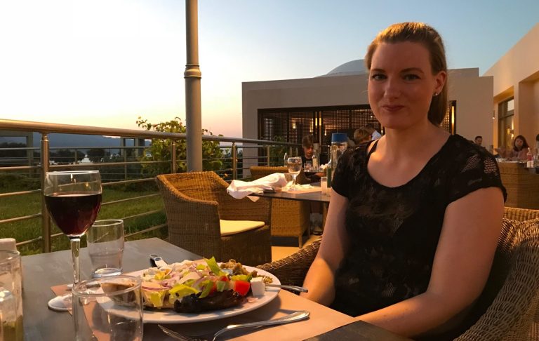 Kvinna sitter vid ett bord med mat och vin, solnedgång i bakgrunden.