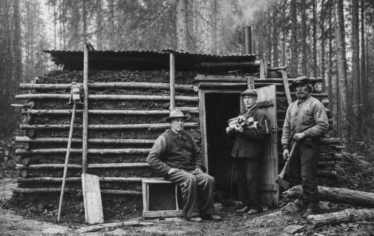 Tre män står vid en enkel träkoja i skogen, en man sitter på marken.