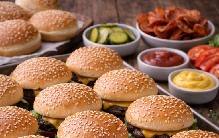 Sesambröd med hamburgare, inlagda gurkor, bacon och såser på träyta.