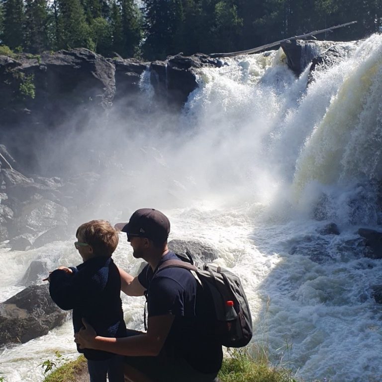 Man och barn står vid en vattenfall, med kraftigt vatten som sprutar omkring dem. 