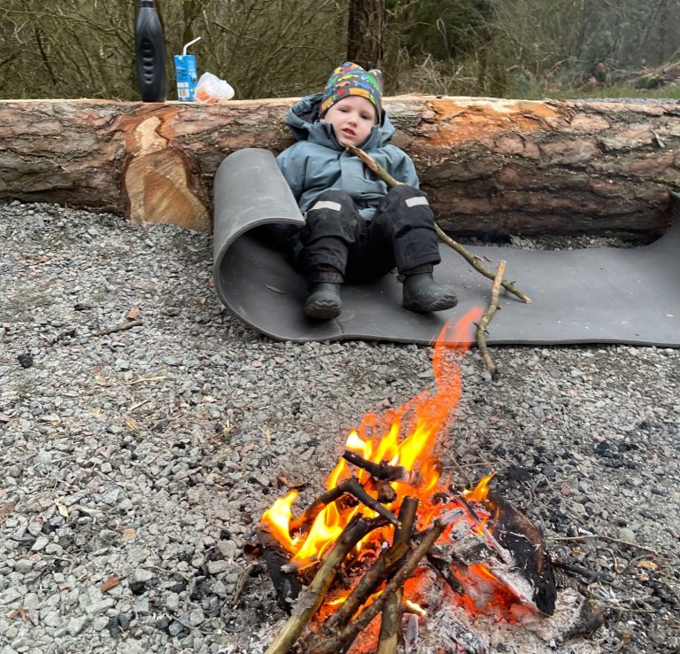 Ett barn sitter på en matta vid en lägereld i skogen.