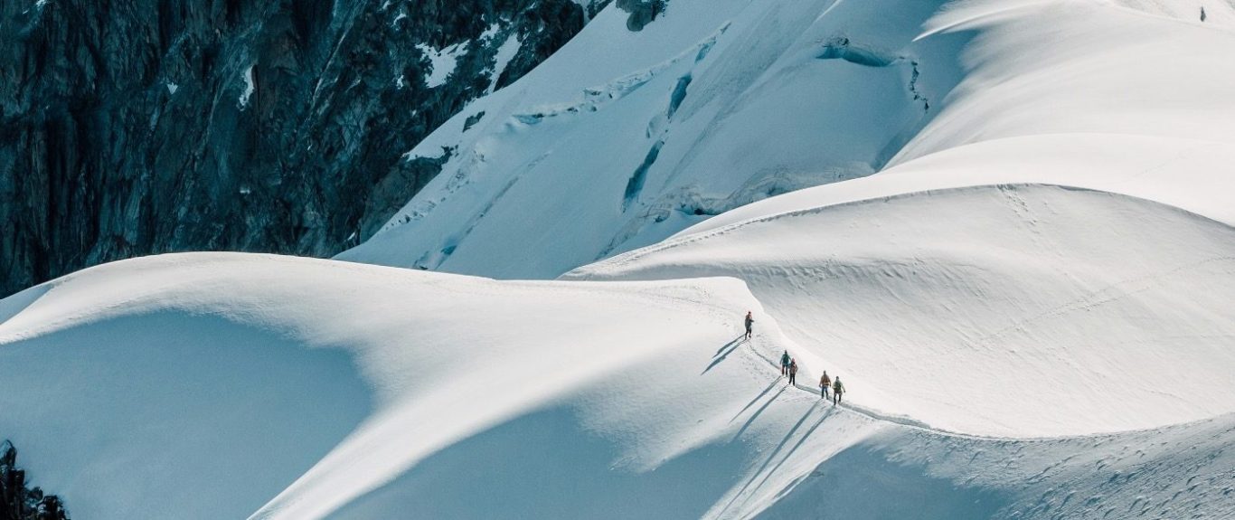 Snötäckta bergstoppar med mjuka kurvor och några människor som vandrar på snön.