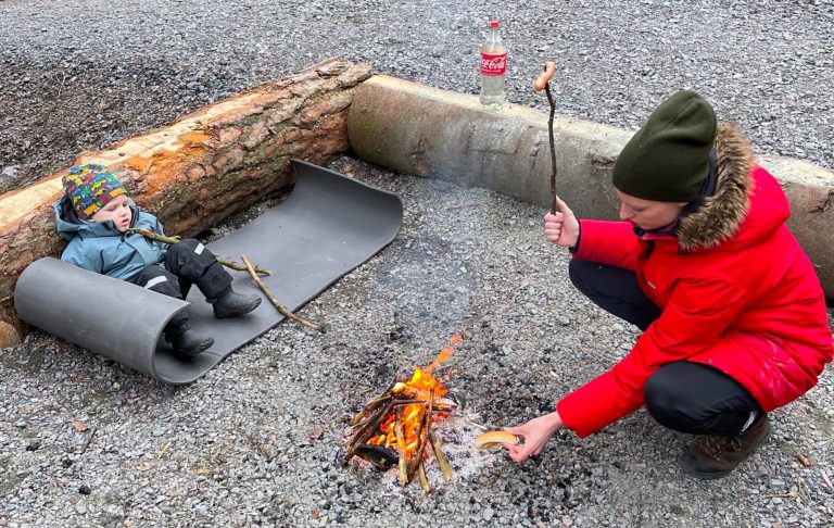 Barn ligger på en matta vid en lägereld, medan en vuxen grillar korv.