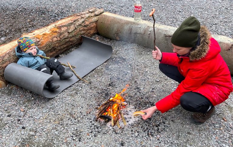 Barn ligger på marken vid en brasa där en vuxen grillar korv.