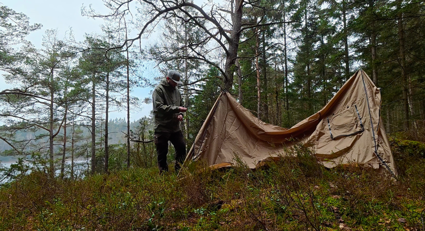 Sätter upp tältet En person slår upp ett tält i en skog omgiven av träd och en sjö i bakgrunden.