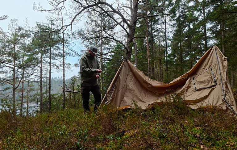 En person står nära ett tält i en skog, omgiven av träd och vatten.