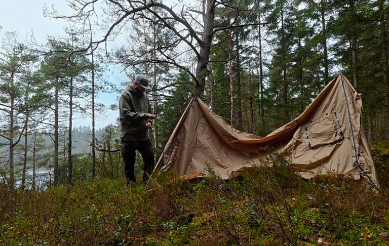 Person står vid ett tält i en skog med träd och en sjö i bakgrunden.