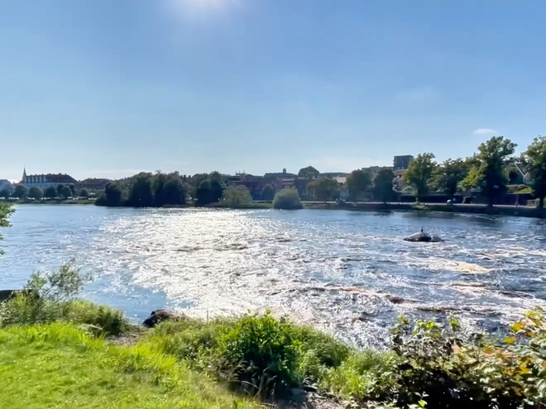 Lugn flod med grönskande bankar och blå himmel i bakgrunden.