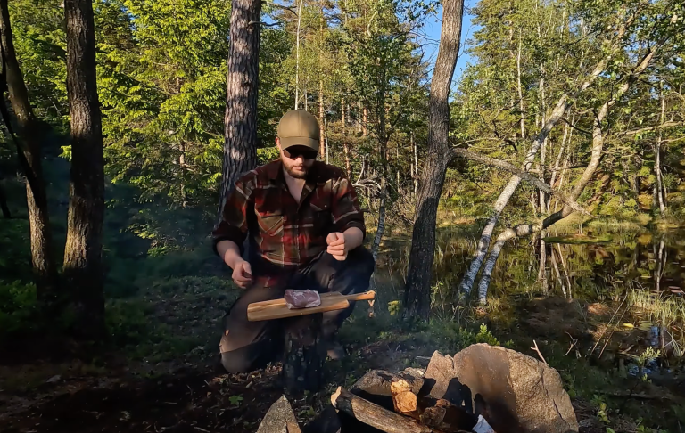 Man lagar mat vid en öppen eld i skogen, omgiven av träd och natur.