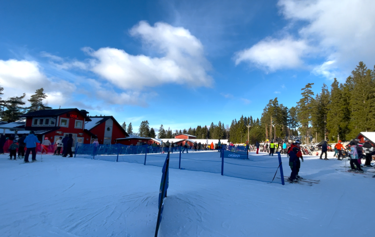 Snöklädd skidort med människor som åker skidor och en röd stuga i bakgrunden.