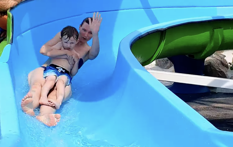 En vuxen och ett barn åker ner för en vattenrutschkana i färgglad miljö.