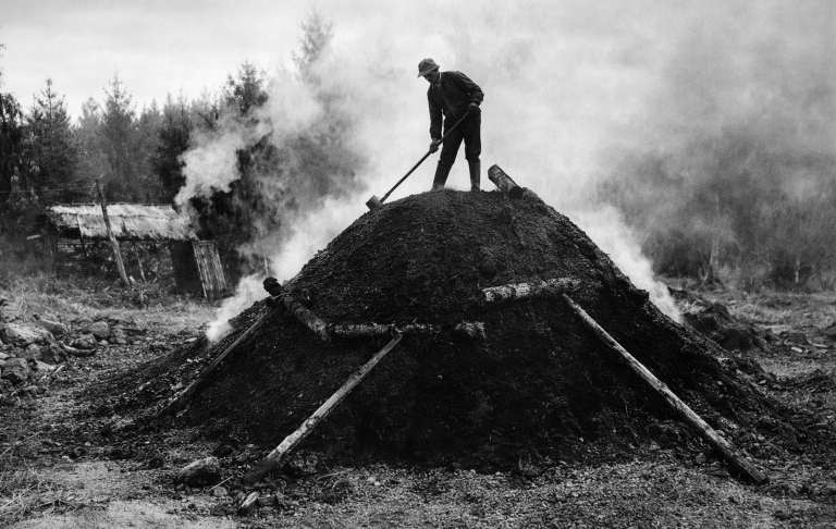 Arbetare på en kolbinge med rök som stiger upp i skogsmiljö.