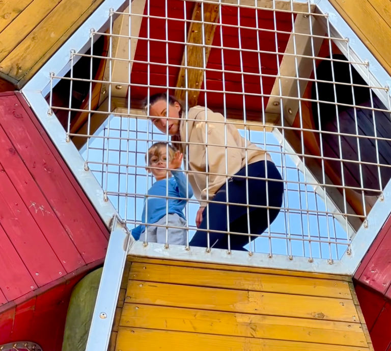 En vuxen och ett barn står på en lekplats under ett färgglatt lekredskap.