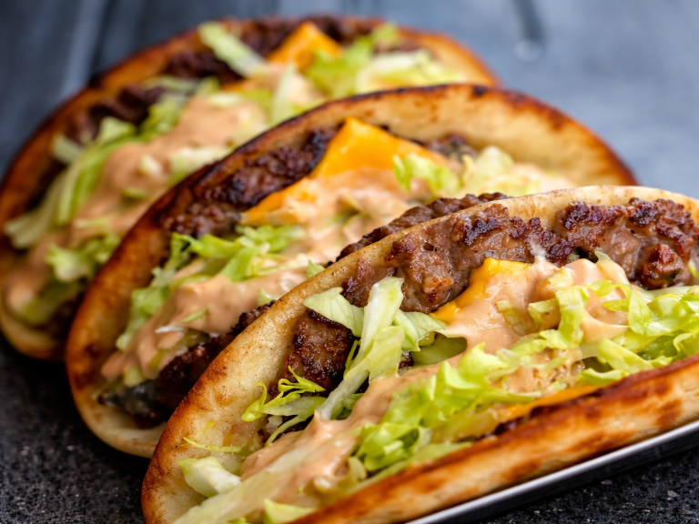 Tre Smashed Taco Burgers  fyllda med kött, sallad och sås, serverade på en mörk bakgrund.