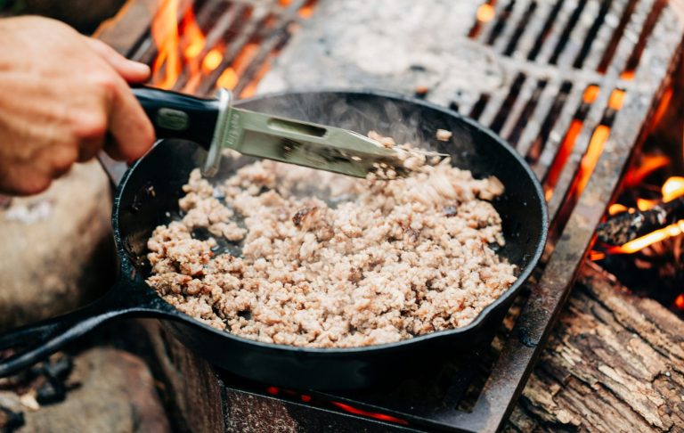 Stekt köttfärs i en stekpanna över öppen eld. Rök och glöd syns runtom.