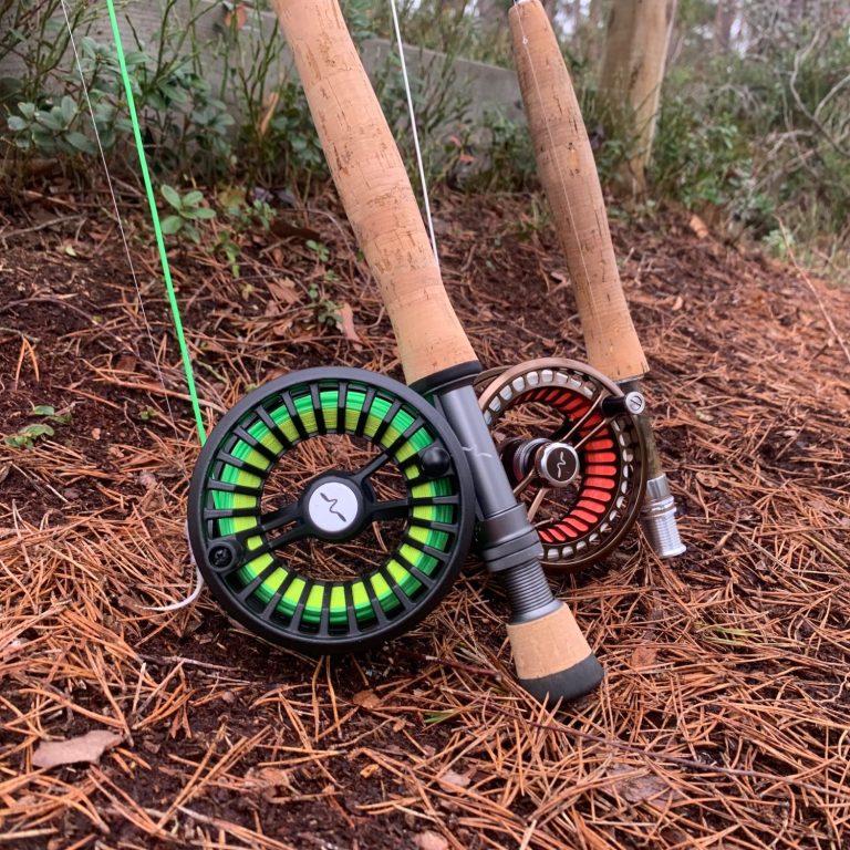 Två flugfiskespön från guideline med rullar i grönt och svart, placerade på barrig mark.