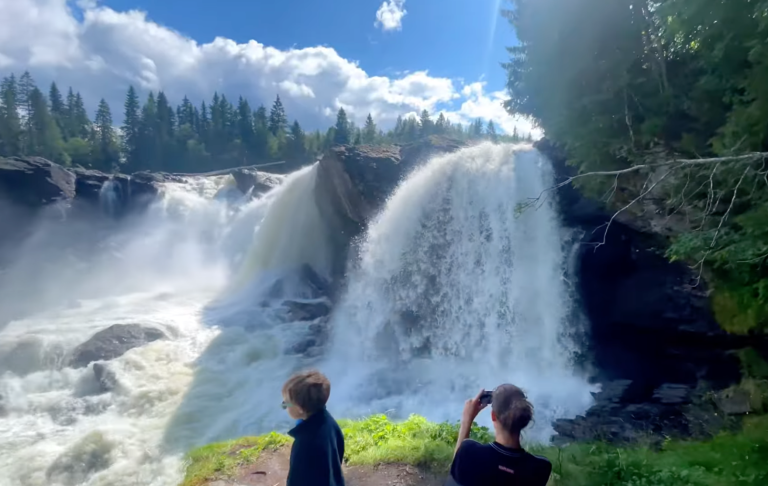 En vattenfall med kraftig ström, omgiven av grönska och klarblå himmel.