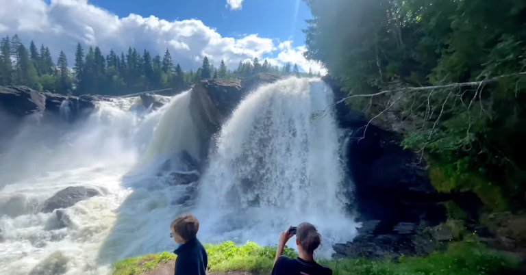 Ett storslaget vattenfall med två personer i förgrunden. Många träd i bakgrunden.