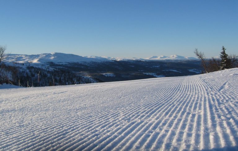 Snötäckt landskap med spår i snön och fjäll i bakgrunden under klarblå himmel.