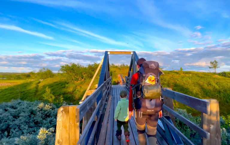 Två personer går över en träbro i en naturskön miljö under en klar himmel.