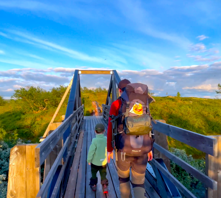 Familj går över bron till Harrsjöstugorna uppe i Fulufjällets Nationalpark i solnedgången.