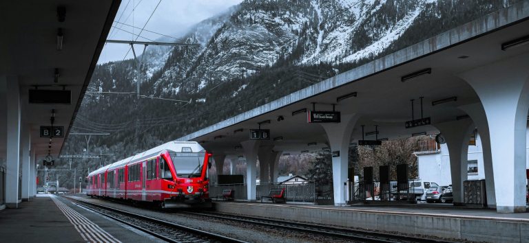 Röd tåg vid en plattform med berg i bakgrunden under en mulen dag.