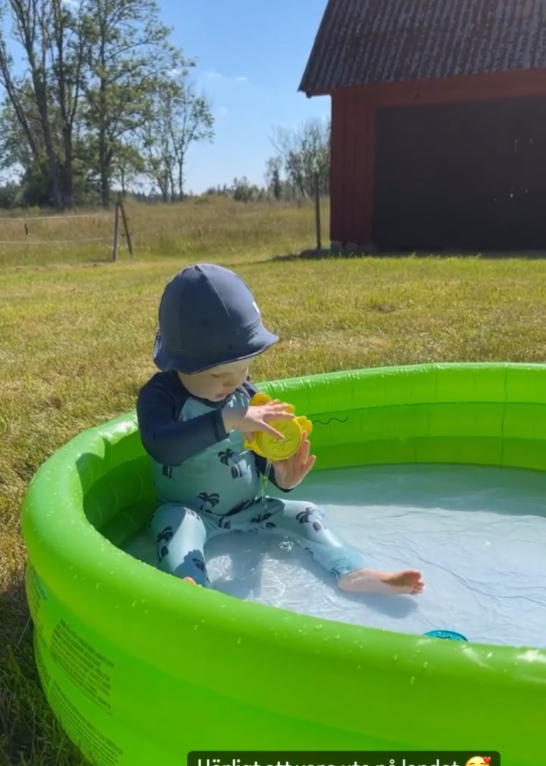 Litet barn i en grön pool på gräset, leker med en leksak i soligt väder.